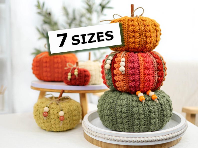 May include: A collection of crocheted pumpkins in various colors including orange, red, green, and cream, featuring textured stitch patterns, wooden bead accents, and brown stems. A white sign with black text reads 7 SIZES, displayed on trays.