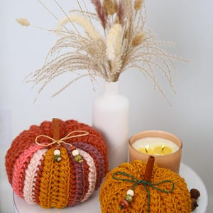 May include: Two crocheted pumpkins featuring ribbed textures in shades of orange, pink, and brown. Each pumpkin is topped with a cinnamon stick stem, a thin twine or string bow, and small wooden beads in green, brown, and cream colors.