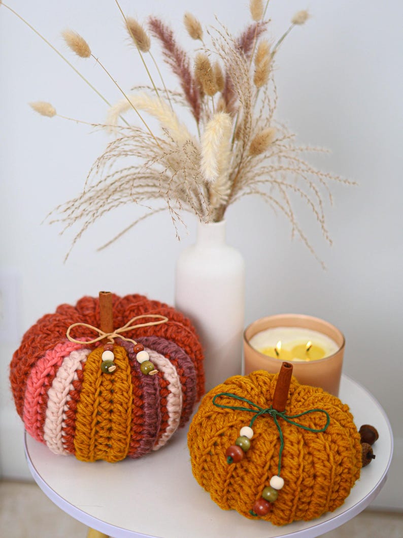 May include: Two crocheted pumpkins featuring ribbed textures in shades of orange, pink, and brown. Each pumpkin is topped with a cinnamon stick stem, a thin twine or string bow, and small wooden beads in green, brown, and cream colors.