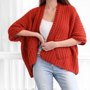 May include: A red crocheted cardigan with a long, open front and 3/4 sleeves. The cardigan has a textured stitch pattern and a decorative trim around the neckline and sleeves.