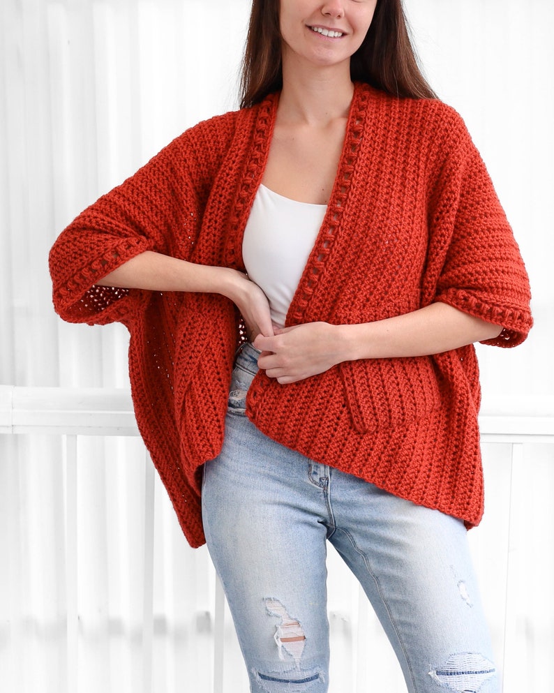 May include: A red crocheted cardigan with a long, open front and 3/4 sleeves. The cardigan has a textured stitch pattern and a decorative trim around the neckline and sleeves.