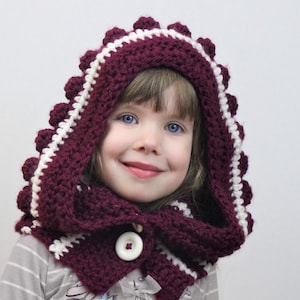 May include: A burgundy crocheted hooded scarf with white trim and pom-poms. The scarf has a button closure and is worn by a young girl.