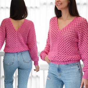 May include: A pink crocheted sweater with a deep V-neck and long sleeves. The sweater has a loose fit and a delicate openwork pattern.