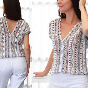 Puede incluir: Top de crochet de manga corta con cuello en V en una paleta de colores neutros de blanco, gris y marr&oacute;n. El top presenta un borde festoneado a lo largo del escote y las mangas. El dise&ntilde;o de tejido abierto es visible.