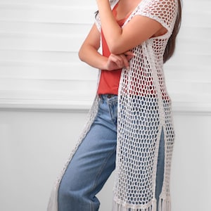 May include: A long white crochet vest featuring an open-knit mesh pattern, short sleeves, and side slits. The hem is finished with long, straight fringe detailing. The garment is worn over a rust-colored camisole and light blue denim jeans.