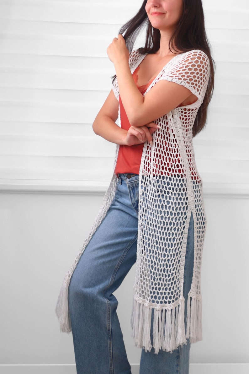May include: A long white crochet vest featuring an open-knit mesh pattern, short sleeves, and side slits. The hem is finished with long, straight fringe detailing. The garment is worn over a rust-colored camisole and light blue denim jeans.