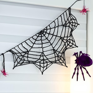 May include: A black crocheted spider web decoration featuring two small pink glittery spiders attached to the ends of the web and one large purple spider positioned separately to the side of the web structure.