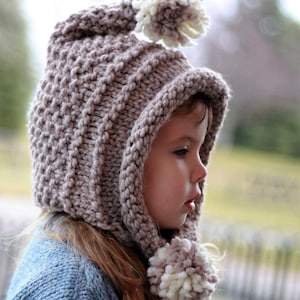 May include: A little girl wearing a brown knitted hooded hat with a white pom-pom on top. The hat has a long, pointed hood and a matching pom-pom on the end of the hood.