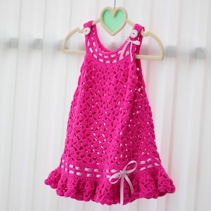 May include: A pink crocheted baby dress with white ribbon accents and white buttons. The dress has a scalloped hem and is hanging on a wooden hanger.