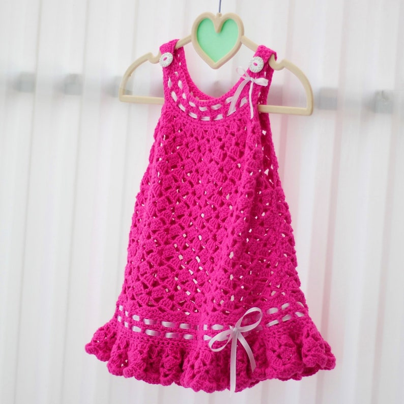 May include: A pink crocheted baby dress with white ribbon accents and white buttons. The dress has a scalloped hem and is hanging on a wooden hanger.