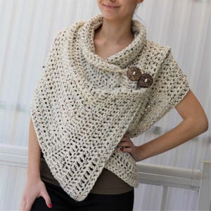 May include: A cream-coloured crocheted shawl with a cowl neck and wooden button fastening. The shawl is draped over a person's shoulders and is worn with a brown top and black trousers.