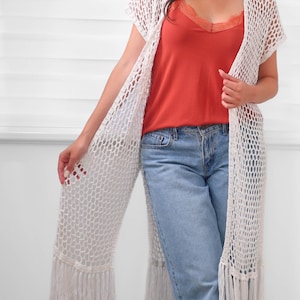 May include: A long, white, open-front crochet cardigan featuring a loose, net-like stitch pattern and straight fringe trim along the bottom hem. The garment is layered over a solid orange camisole with lace detailing and light blue denim jeans.