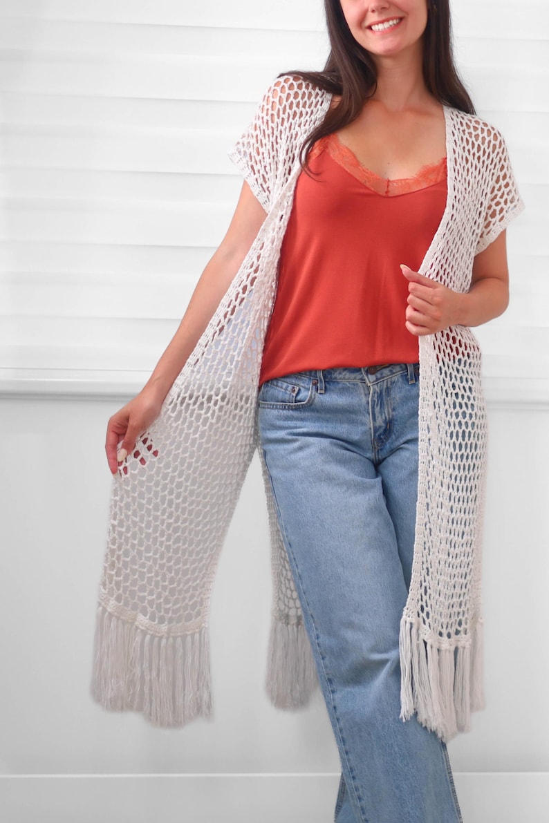 May include: A long, white, open-front crochet cardigan featuring a loose, net-like stitch pattern and straight fringe trim along the bottom hem. The garment is layered over a solid orange camisole with lace detailing and light blue denim jeans.
