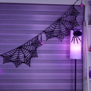 May include: A black crocheted spider web garland featuring small pink LED spider lights. The garland is draped horizontally, with a large black spider attached to a tall floor lamp that emits a soft purple glow, creating a dark atmosphere.