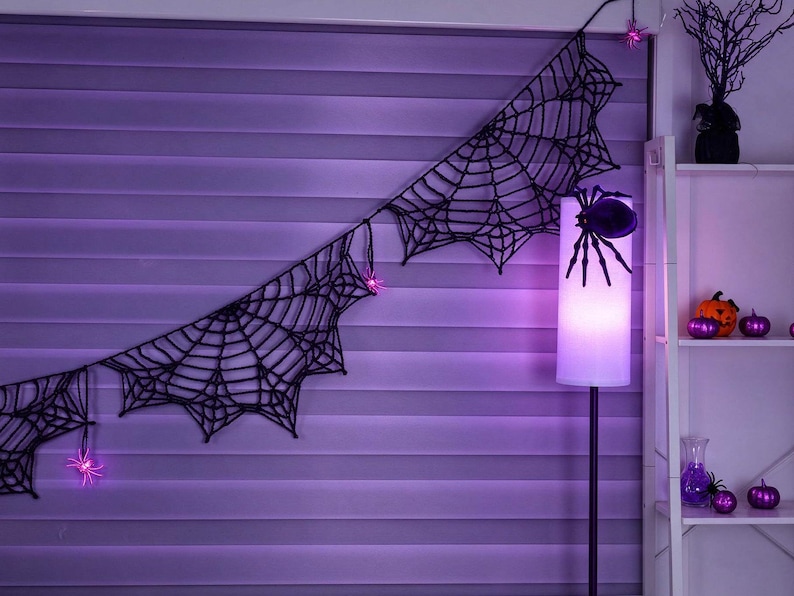 May include: A black crocheted spider web garland featuring small pink LED spider lights. The garland is draped horizontally, with a large black spider attached to a tall floor lamp that emits a soft purple glow, creating a dark atmosphere.