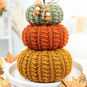 May include: A stack of three crocheted pumpkins in shades of green, orange, and yellow. The top pumpkin is green with wooden beads and a twine stem. The middle pumpkin is orange, and the bottom pumpkin is yellow. The pumpkins are stacked on a white tray.