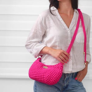 May include: A vibrant hot pink crochet handbag featuring a textured, knotted stitch pattern. The bag includes a long, matching pink cord strap with gold-toned metal end caps and a small, oval-shaped metallic brand tag attached to the front center.