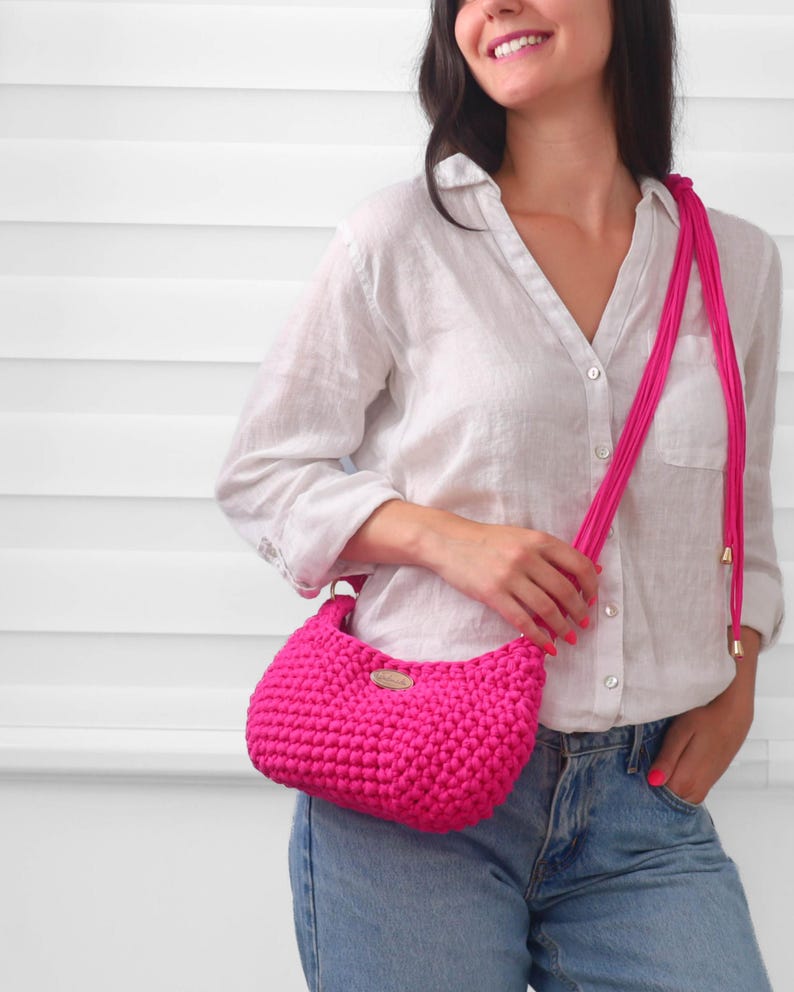 May include: A vibrant hot pink crochet handbag featuring a textured, knotted stitch pattern. The bag includes a long, matching pink cord strap with gold-toned metal end caps and a small, oval-shaped metallic brand tag attached to the front center.