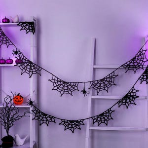 May include: A black crocheted spider web garland featuring multiple web segments and small dangling spiders, draped across a white ladder shelf. The garland is composed of interconnected web patterns with thin yarn strands.