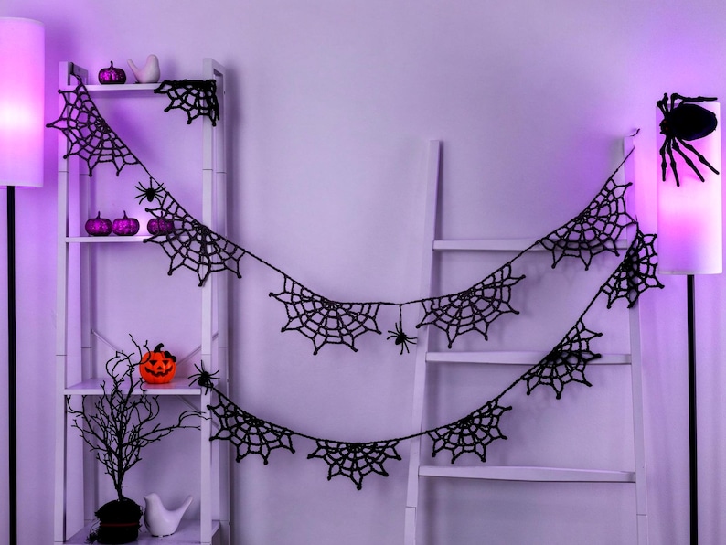 May include: A black crocheted spider web garland featuring multiple web segments and small dangling spiders, draped across a white ladder shelf. The garland is composed of interconnected web patterns with thin yarn strands.