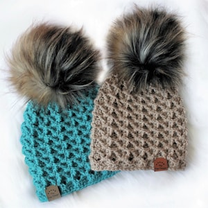 May include: Two crocheted hats with faux fur pom poms. The hats are in a teal and brown color. The hats have a label that says "The Easy Design".