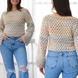 May include: A cream-colored, open-weave crochet sweater with long, flared sleeves. The sweater has a scoop neckline and a subtle color gradient of peach, blue, and gray. The person is wearing light blue ripped jeans.