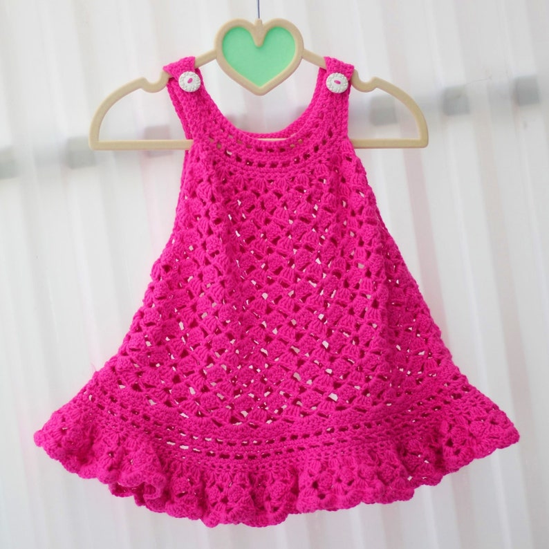 May include: A pink crocheted baby dress with a ruffled hem and white buttons.