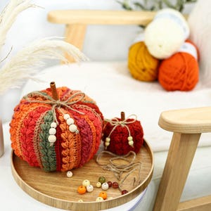 May include: Two crocheted pumpkins in shades of orange, red, and green, featuring a cinnamon stick stem and twine bow with wooden beads. The pumpkins sit on a round wooden tray alongside loose wooden beads and a length of twine.