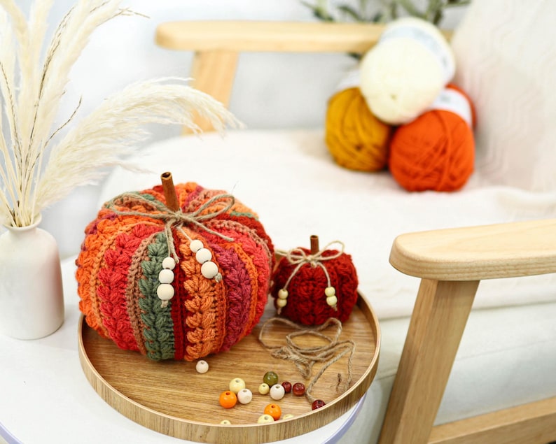 May include: Two crocheted pumpkins in shades of orange, red, and green, featuring a cinnamon stick stem and twine bow with wooden beads. The pumpkins sit on a round wooden tray alongside loose wooden beads and a length of twine.