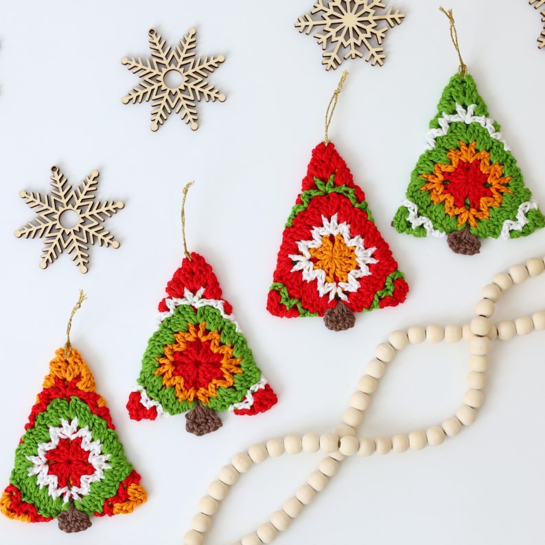 Puede incluir: Cuatro adornos de &aacute;rbol de Navidad de ganchillo en rojo, verde, blanco y naranja con una base de fieltro marr&oacute;n. Los adornos cuelgan de una cuerda blanca y se muestran sobre una superficie blanca con copos de nieve de madera y una cadena de cuentas de madera.