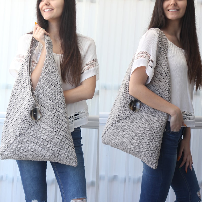 Op de afbeelding: Een grijze gehaakte tote bag met een grote, ronde, bruine knoop. De tas wordt over de schouder gedragen en heeft een nonchalante, casual look.