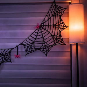 May include: A black crocheted spider web decoration featuring two small, bright pink plastic spiders attached to the yarn structure. The web is displayed against horizontal window blinds, with a warm-toned floor lamp nearby.