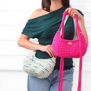 May include: Two crochet handbags featuring a textured, rounded silhouette. One bag is a vibrant solid hot pink with long, dangling cord fringe and a small oval tag. The second bag is a light gray with flecks of green and blue, also with a small tag.