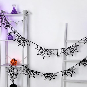 May include: A black crocheted spider web garland featuring multiple web segments connected by a thin string, accented with small black spider figures attached to the webs. The garland is draped across a white ladder-style shelving unit.