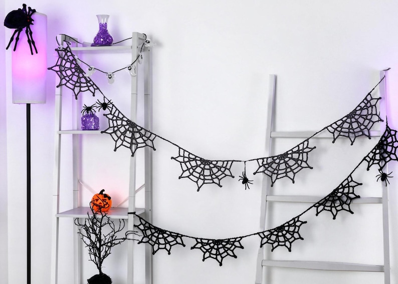 May include: A black crocheted spider web garland featuring multiple web segments connected by a thin string, accented with small black spider figures attached to the webs. The garland is draped across a white ladder-style shelving unit.