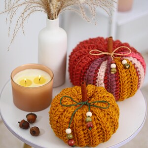 May include: Two crocheted pumpkins in mustard yellow and multi-colored yarn, each topped with a cinnamon stick stem. The pumpkins are decorated with thin twine bows and small wooden beads. Three small brown acorns sit nearby on the surface.