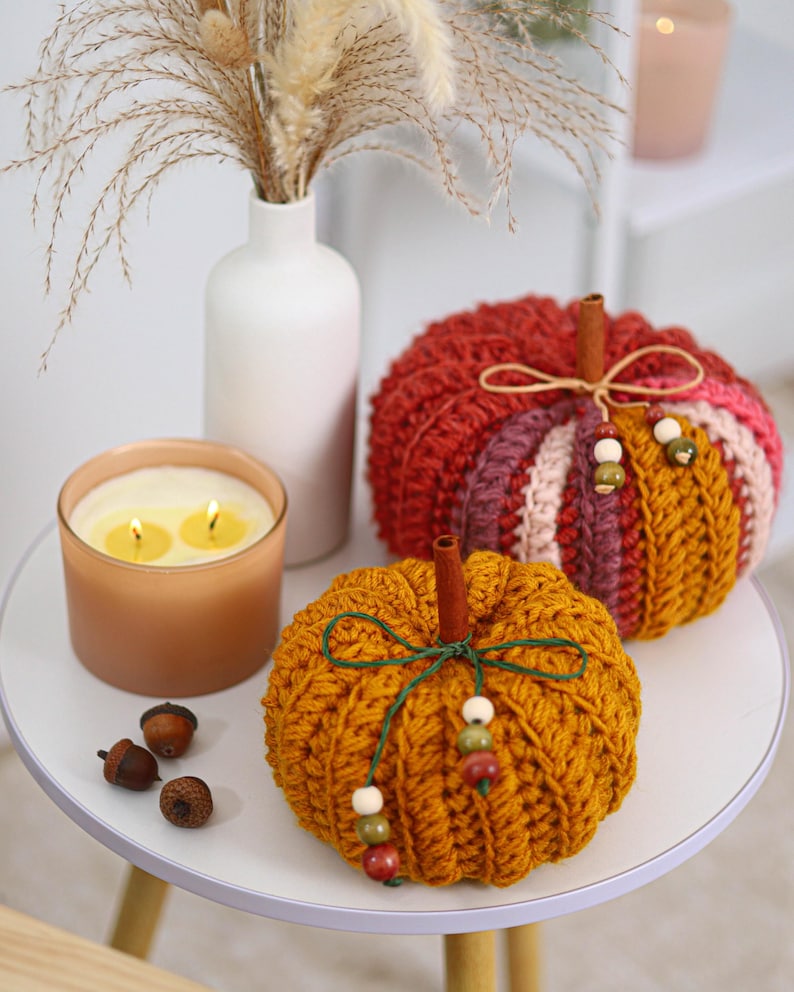 May include: Two crocheted pumpkins in mustard yellow and multi-colored yarn, each topped with a cinnamon stick stem. The pumpkins are decorated with thin twine bows and small wooden beads. Three small brown acorns sit nearby on the surface.