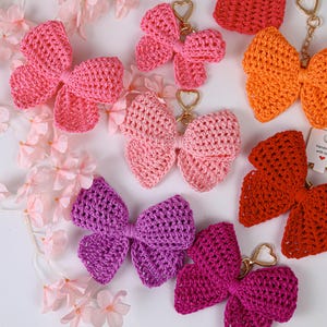 May include: A collection of seven crocheted bow keychains in various colors including pink, orange, red, purple, and magenta. Each bow features a gold-toned heart-shaped clasp. One red bow has a small white tag with the text Handmade with love.