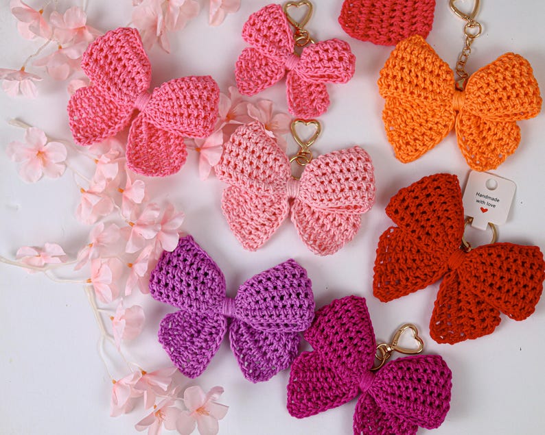 May include: A collection of seven crocheted bow keychains in various colors including pink, orange, red, purple, and magenta. Each bow features a gold-toned heart-shaped clasp. One red bow has a small white tag with the text Handmade with love.