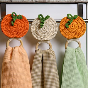 May include: Three hanging kitchen towels featuring crocheted pumpkin toppers in orange and cream with green stems. Each towel is attached to a light-colored wooden ring. The towels have waffle-weave textures in peach, tan gingham, and light green.