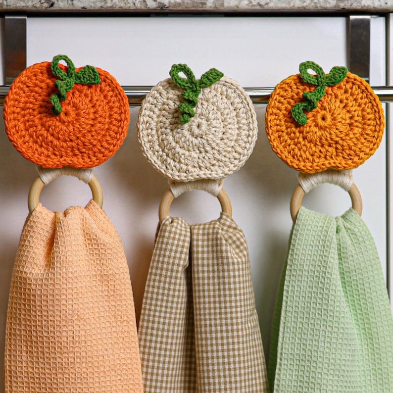 May include: Three hanging kitchen towels featuring crocheted pumpkin toppers in orange and cream with green stems. Each towel is attached to a light-colored wooden ring. The towels have waffle-weave textures in peach, tan gingham, and light green.