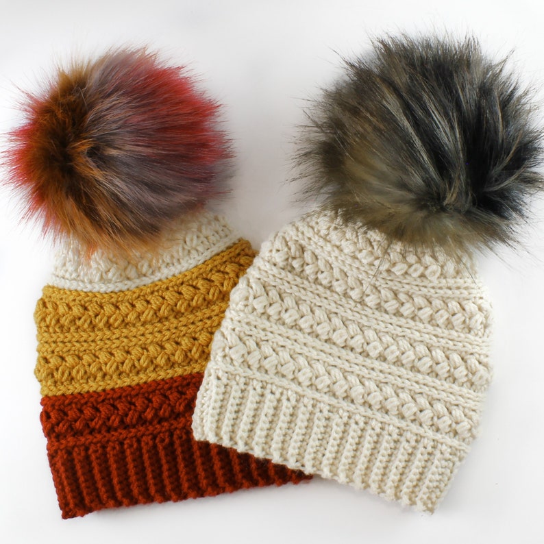 Puede incluir: Dos gorros de punto con pompones de piel sint&eacute;tica. Un gorro es crema con un pompon marr&oacute;n, el otro es multicolor con un pompon marr&oacute;n, rojo y gris.