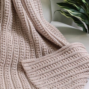 May include: A soft, beige crocheted throw blanket with a textured pattern. The blanket is folded and draped over a chair.