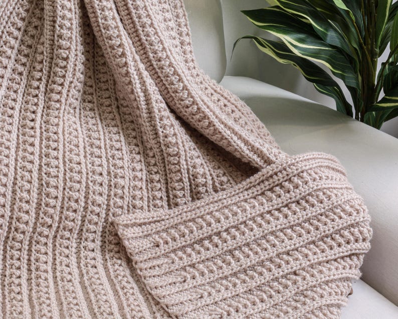 May include: A soft, beige crocheted throw blanket with a textured pattern. The blanket is folded and draped over a chair.