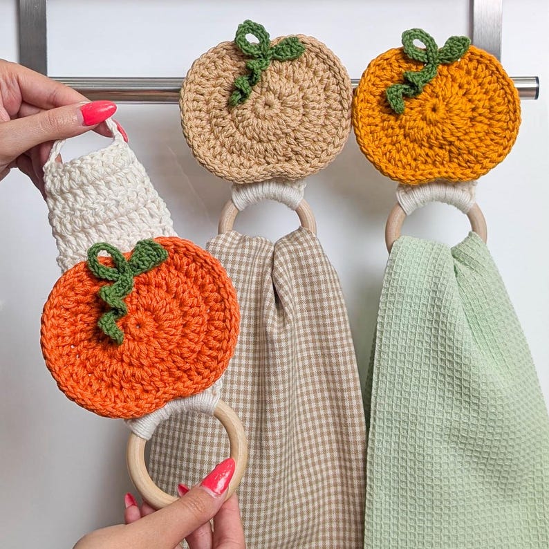 May include: Three crochet pumpkin towel holders featuring orange and beige yarn pumpkins with green stems. Each pumpkin is attached to a wooden ring that holds a hanging kitchen towel in either a beige gingham or a solid light green waffle weave.