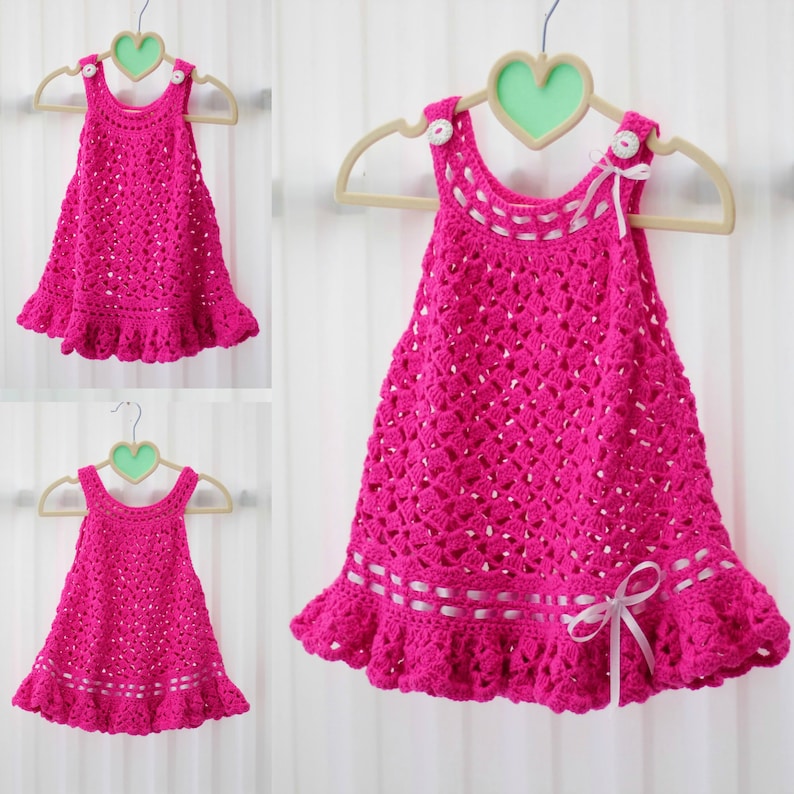 May include: A pink crocheted baby dress with white ribbon accents and white buttons. The dress has a scalloped hem and is hanging on a white hanger.