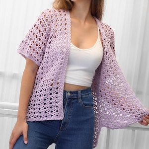May include: A light purple crochet cardigan with a lacy open stitch pattern. The cardigan has short sleeves and a relaxed fit.