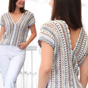 May include: A short-sleeved, V-neck crochet top in neutral tones of white, grey, and beige. The open-weave design creates a textured appearance. The top is a handmade garment, suitable for casual wear.