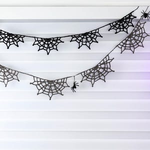 May include: A black crochet garland featuring multiple spider web motifs connected by a thin string. Small black spider figures are attached to the web patterns at various points along the length of the decorative hanging piece.