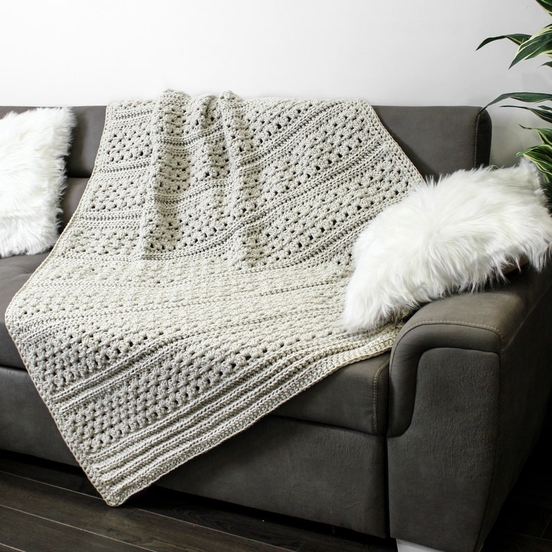 May include: A beige crocheted throw blanket draped over a gray couch with a white faux fur pillow.